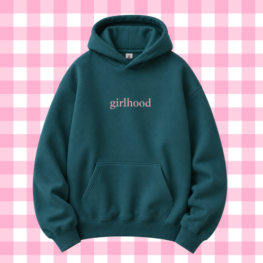 A dark teal-green pullover hoodie with the word 'girlhood' printed in a light pink serif font on the chest, displayed against a pink and white checkered background.