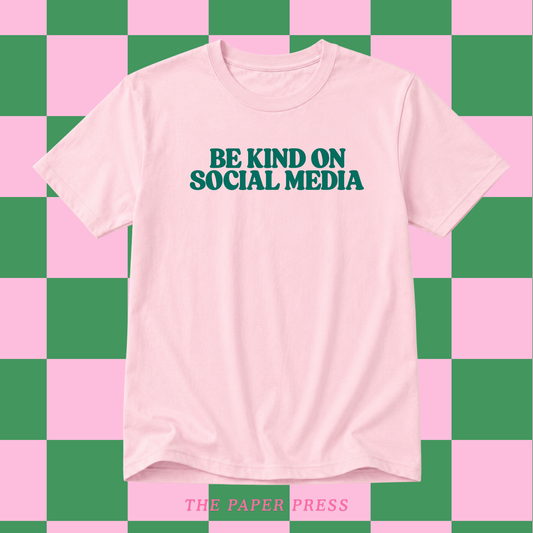 Baby pink slogan t-shirt that says be kind on social media in retro dark green font.