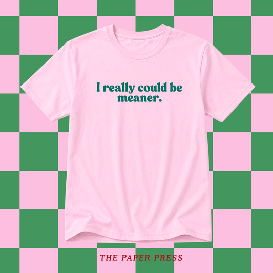 A light pink short-sleeve t-shirt with the text 'I really could be meaner.' printed in dark teal across the chest, displayed against a pink and green checkered background.