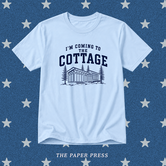 A light blue crewneck t-shirt with a dark navy graphic that reads 'I'M COMING TO THE COTTAGE' above an illustration of a cottage and pine trees. The shirt is displayed flat against a textured blue background patterned with white stars, with 'THE PAPER PRESS' printed at the bottom.