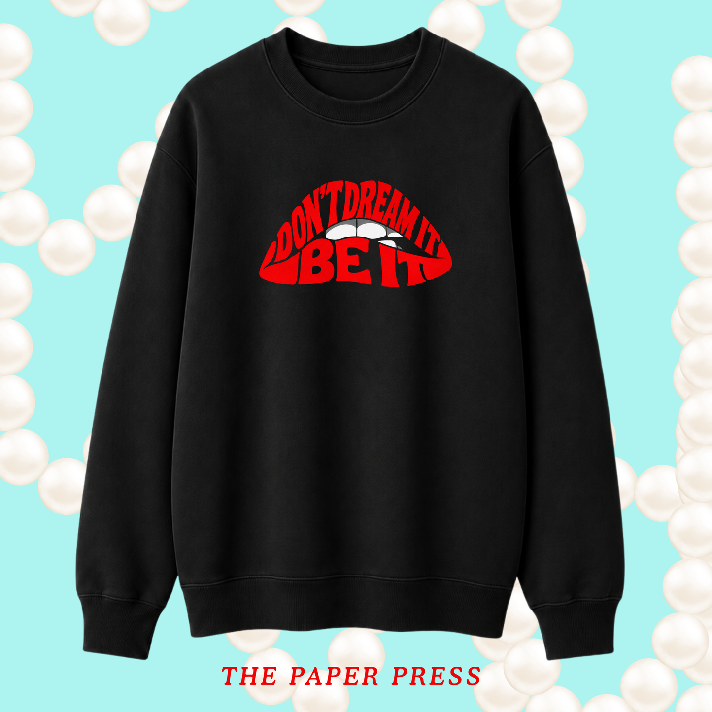 A black crewneck sweatshirt with a red graphic on the chest reading 'I DON'T DREAM IT BE IT' over white lips, displayed on a light blue background with white pearl-like circles and the text 'THE PAPER PRESS' at the bottom.