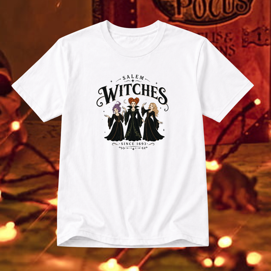 A white short-sleeve graphic t-shirt featuring the 'Salem Witches' illustration, displayed against a warm, blurred Halloween-themed background with orange lights and a cat.