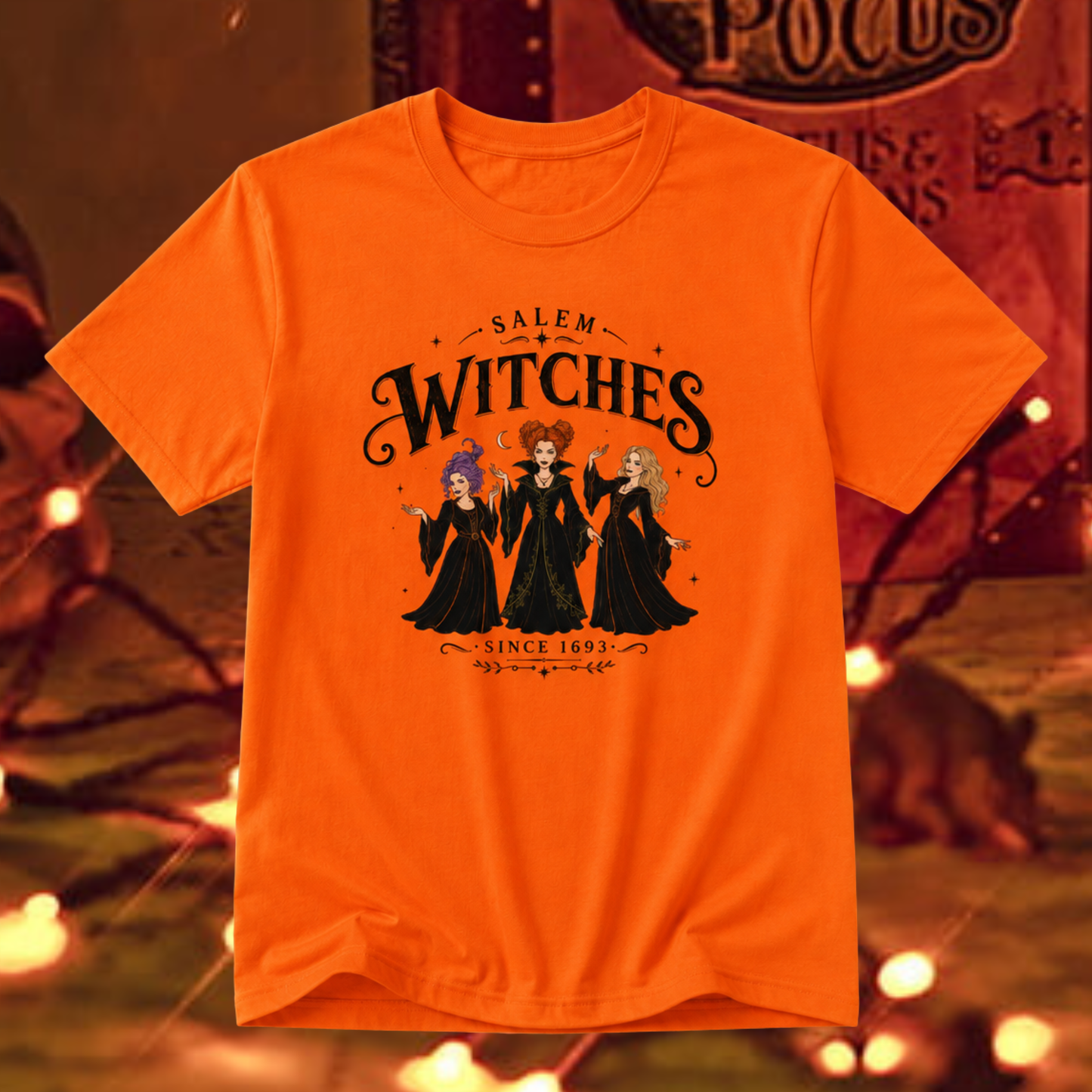 An orange short-sleeve graphic t-shirt featuring the characters 'Salem Witches' in black robes with colorful hair, displayed against a warm, blurred background with warm lights.