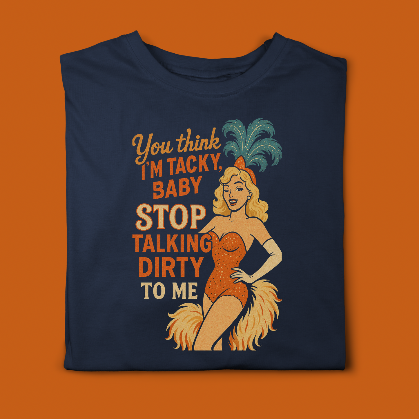 Folded navy blue T-shirt featuring a vintage showgirl graphic with the slogan ‘You think I’m tacky, baby, stop talking dirty to me’ in orange and cream lettering. The blonde showgirl wears an orange sequined outfit with feather details and a teal feathered headdress.