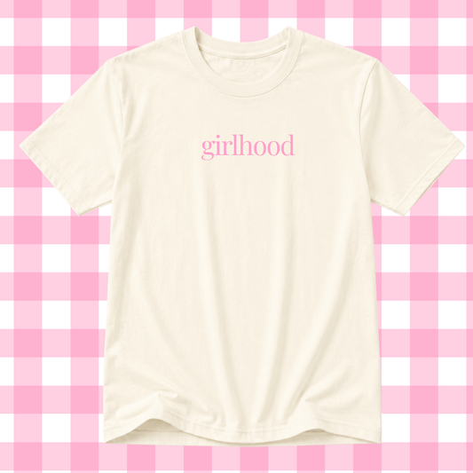An off-white short-sleeve t-shirt with the word 'girlhood' printed in pink lowercase serif letters across the chest, displayed against a pink and white checkered background.