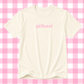 An off-white short-sleeve t-shirt with the word 'girlhood' printed in pink lowercase serif letters across the chest, displayed against a pink and white checkered background.