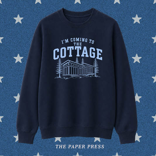 A navy blue crewneck sweatshirt with a light blue graphic print that reads 'I'M COMING TO THE COTTAGE' above an illustration of a cabin and trees. The sweatshirt is displayed against a textured blue background with white stars, and 'THE PAPER PRESS' is printed at the bottom.