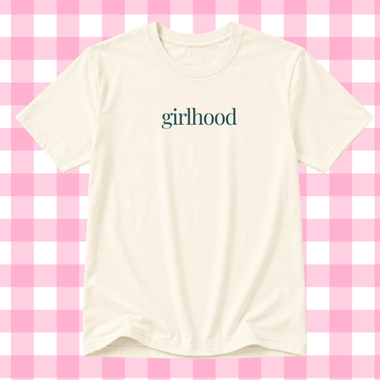 An off-white short-sleeve t-shirt with the word 'girlhood' printed in a dark teal serif font across the chest, displayed against a pink and white checkered background.