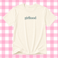 An off-white short-sleeve t-shirt with the word 'girlhood' printed in a dark teal serif font across the chest, displayed against a pink and white checkered background.