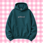 A dark teal-green pullover hoodie with the word 'girlhood' printed in a light pink serif font on the chest, displayed against a pink and white checkered background.
