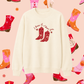 White sweatshirt with cowboy boot design and text on a pink background with colorful boot patterns.