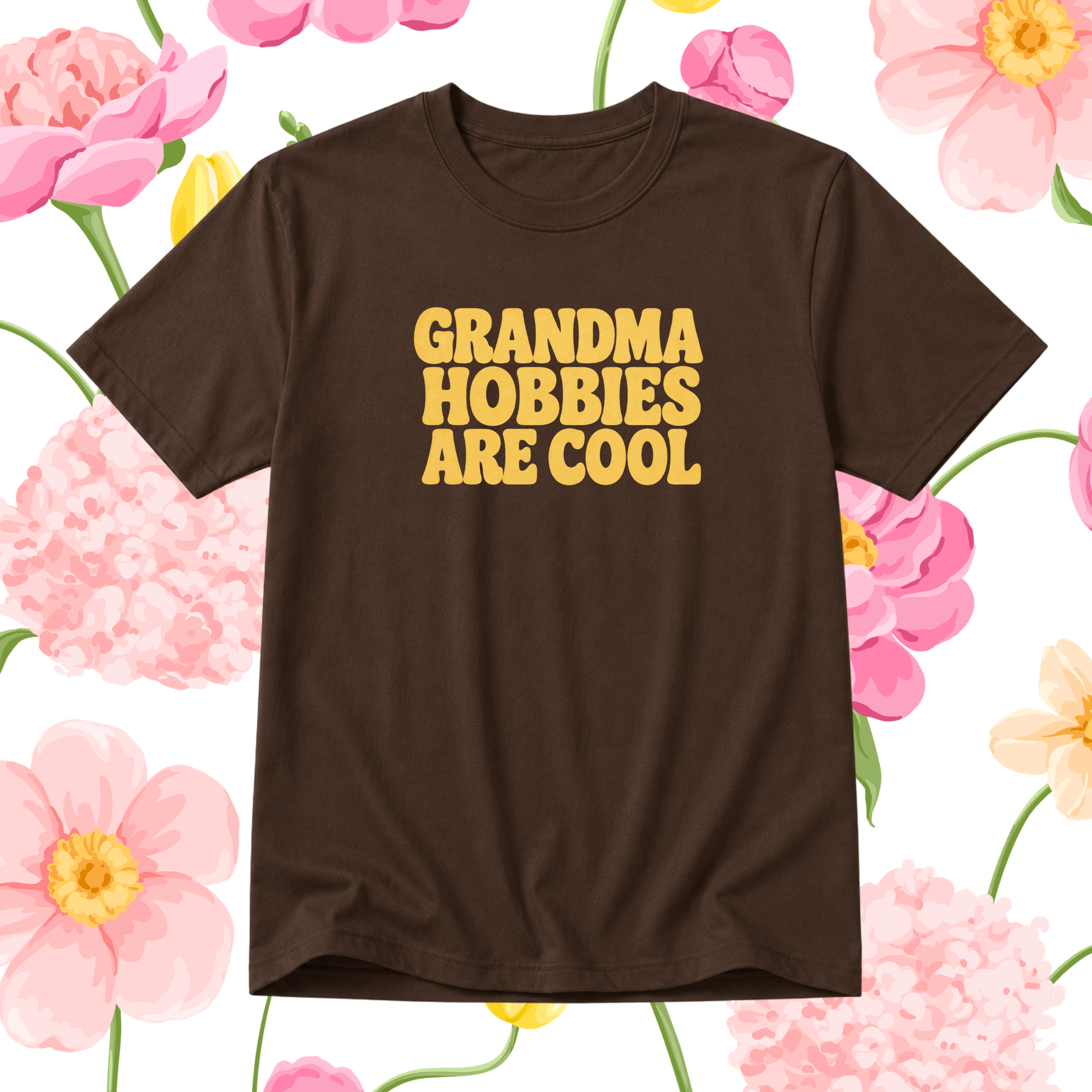 A dark brown short-sleeve t-shirt with the phrase 'GRANDMA HOBBIES ARE COOL' printed in bold yellow letters on the chest, presented against a white background decorated with pink and yellow watercolor flowers.