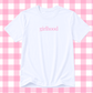A white short-sleeve crewneck t-shirt with the word 'girlhood' printed in pink lowercase serif text across the chest, displayed against a pink and white checkered background.