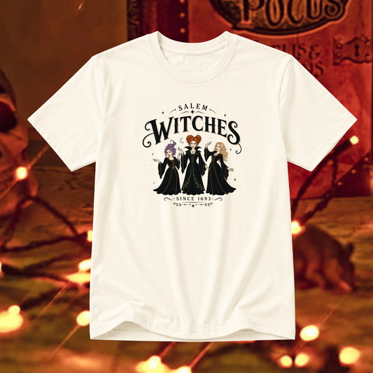 An off-white short-sleeve t-shirt featuring a graphic print of three Salem Witches in black dresses with red, purple, and blonde hair, set against a warm, blurred Halloween-themed background with glowing lights.