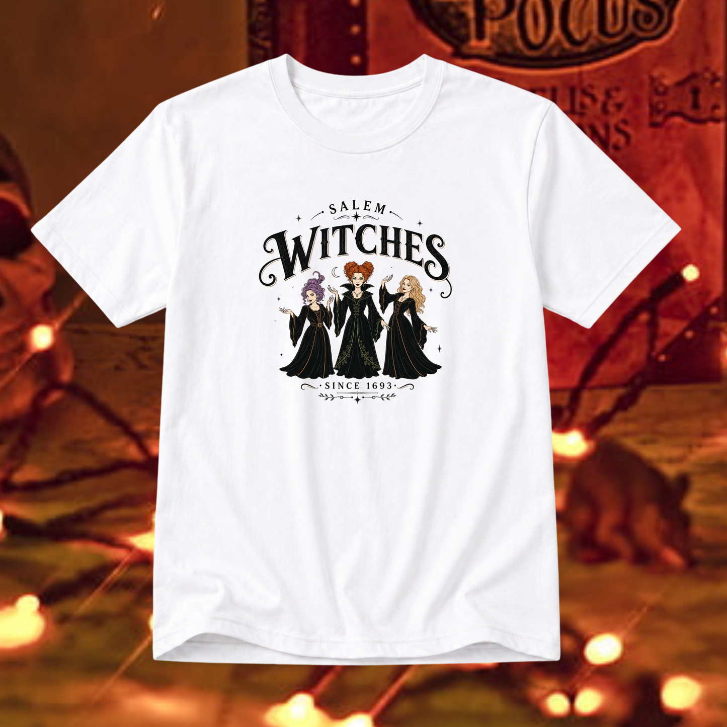 A white short-sleeve graphic t-shirt featuring the 'Salem Witches' illustration, displayed against a warm, blurred Halloween-themed background with orange lights and a cat.