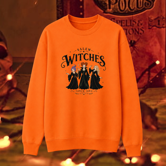 An orange crewneck sweatshirt featuring a graphic print of the Salem Witches, displayed against a warm, atmospheric Halloween-themed background.