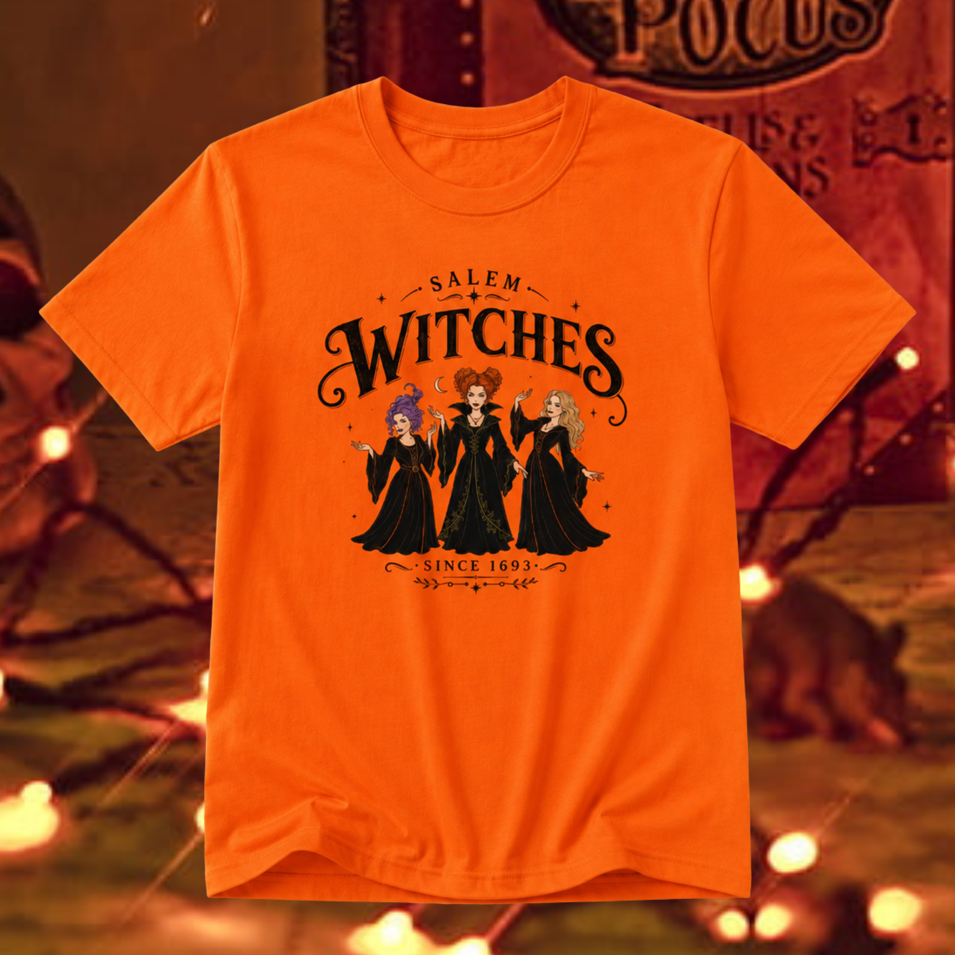 An orange short-sleeve graphic t-shirt featuring the characters 'Salem Witches' in black robes with colorful hair, displayed against a warm, blurred background with warm lights.