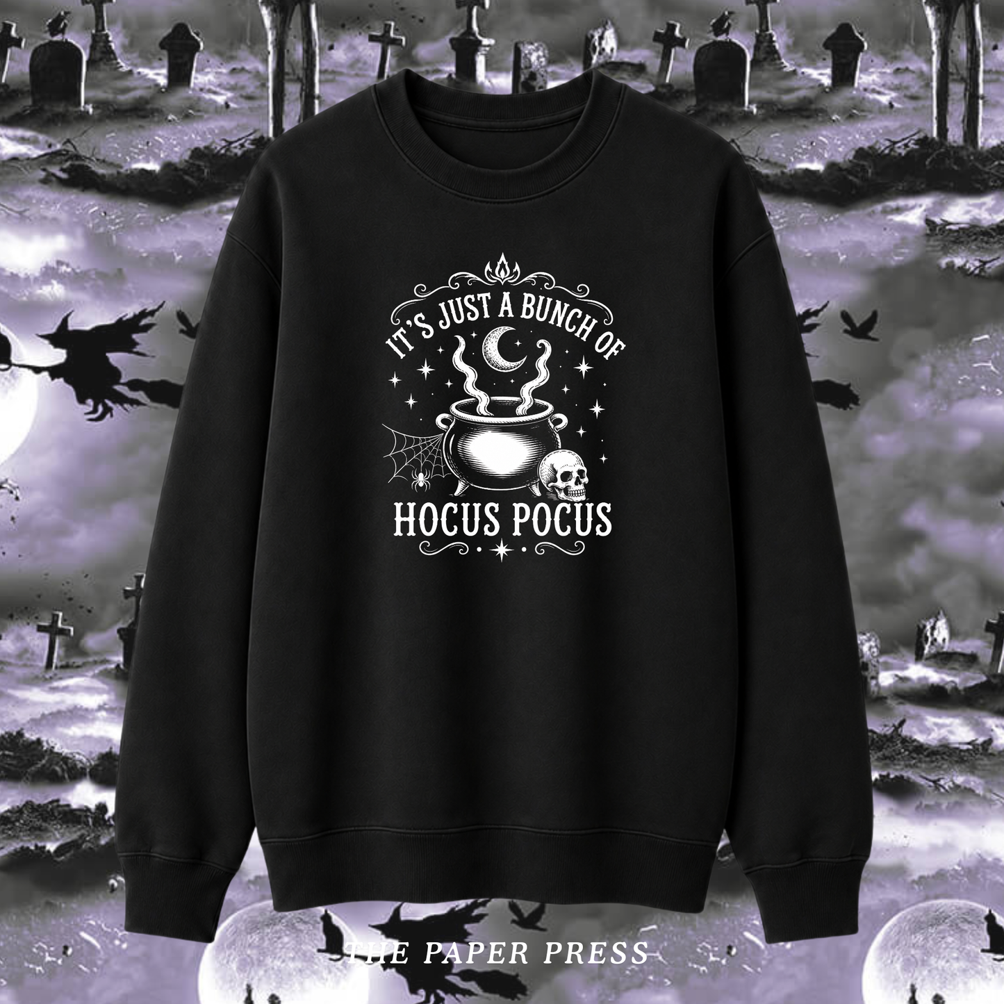 A black crewneck sweatshirt with a white graphic print reading 'IT'S JUST A BUNCH OF HOCUS POCUS' around an illustration of a cauldron, skull, and crescent moon. The sweatshirt is displayed against a spooky Halloween graveyard background with tombstones, crows, and full moons.