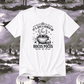 A white short-sleeve crewneck t-shirt with a black graphic print reading 'IT'S JUST A BUNCH OF HOCUS POCUS' around an illustration of a cauldron, skull, spiderwebs, and stars. The shirt is displayed against a spooky, desaturated graveyard background with silhouettes of witches on broomsticks and tombstones.