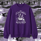 A purple crewneck sweatshirt with a white graphic print reading 'IT'S JUST A BUNCH OF HOCUS POCUS' around an illustration of a cauldron, skull, and stars. The sweatshirt is displayed against a spooky Halloween-themed background of a graveyard with silhouettes of witches flying on broomsticks.