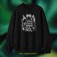 A black crewneck sweatshirt with a white graphic print featuring the text 'oh look another glorious MORNING makes me SICK' surrounded by witch hats, broomsticks, and a cauldron. The sweatshirt is displayed against a dark green and black textured background with 'THE PAPER PRESS' printed in yellow at the bottom.