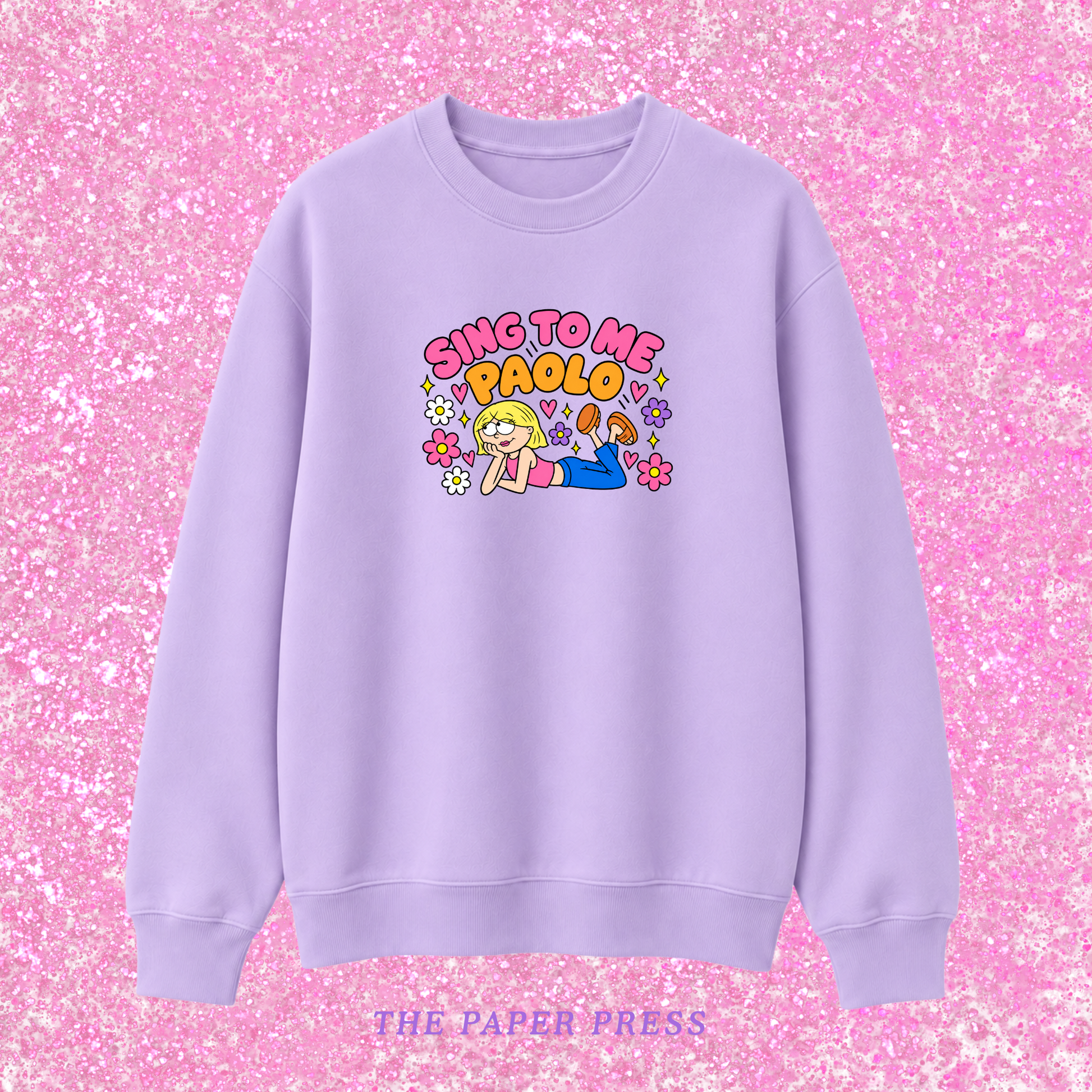 A lavender crewneck sweatshirt featuring a colorful graphic of a cartoon girl and the text 'SING TO ME PAOLO' in pink and yellow, displayed against a pink glitter background with 'THE PAPER PRESS' text at the bottom.