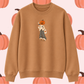 A tan crewneck sweatshirt featuring a central graphic of a scarecrow with a jack-o'-lantern head, set against a light pink background patterned with orange pumpkins.