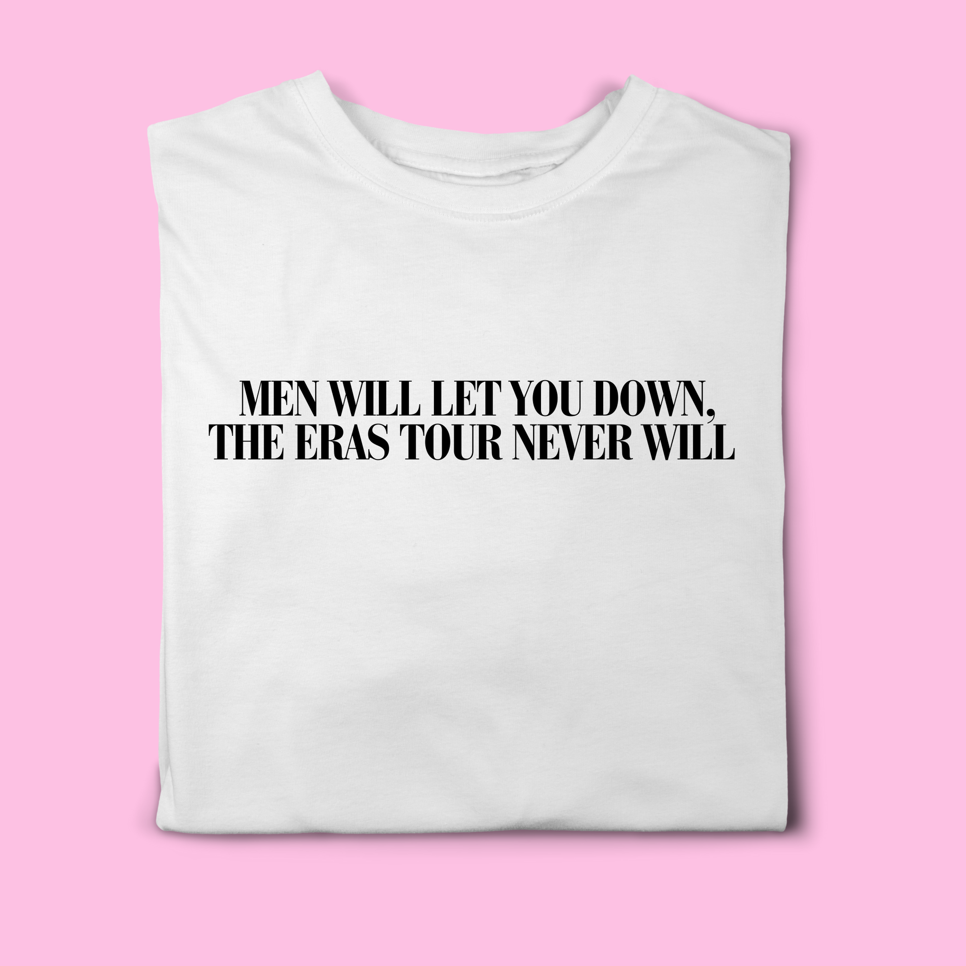 White t-shirt with black text reading “Men Will Let You Down, The Eras Tour Never Will”, folded flat on a pink background